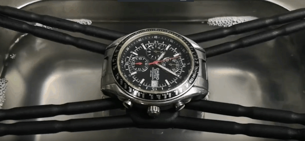 A luxury wristwatch with a metal band suspended in a mesh basket inside an ultrasonic cleaner